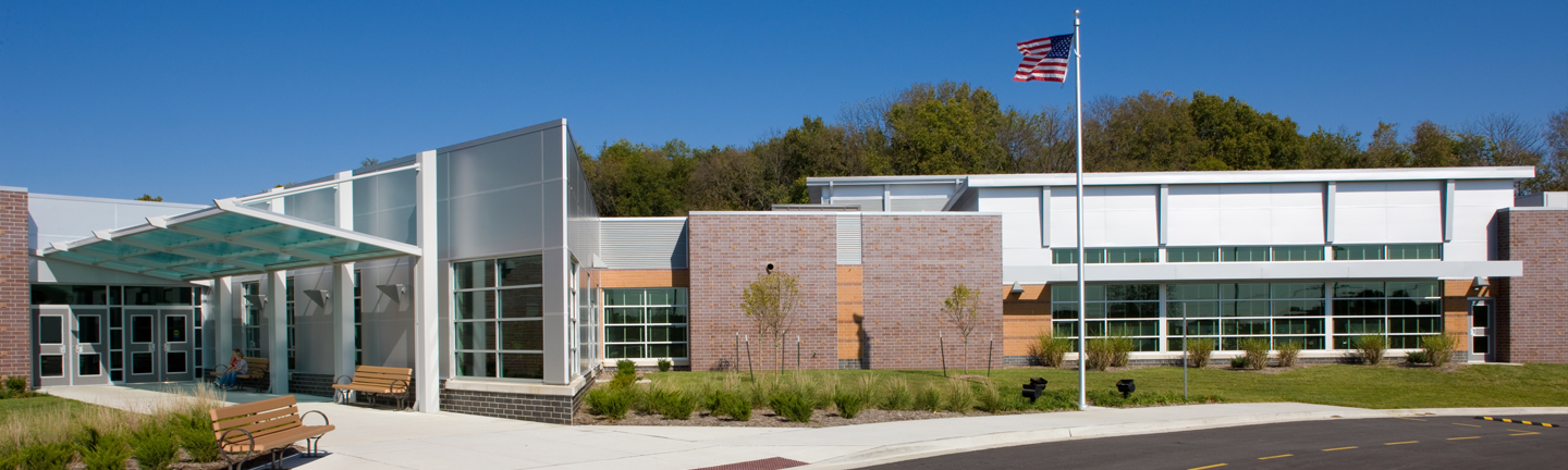 A modern school building with large windows, brick and metal accents, benches along the entrance, landscaped greenery, and an American flag on a tall flagpole under a clear blue sky.