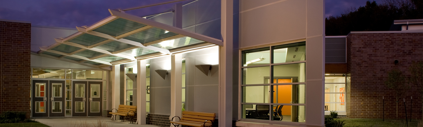A modern building entrance at dusk features glass doors, large windows, illuminated lights, a glass canopy, and benches along the walkway. The sky is darkening, and some interior lights are on.
